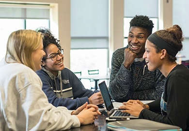Four students smiling while chatting at a table in a residence hall lobby